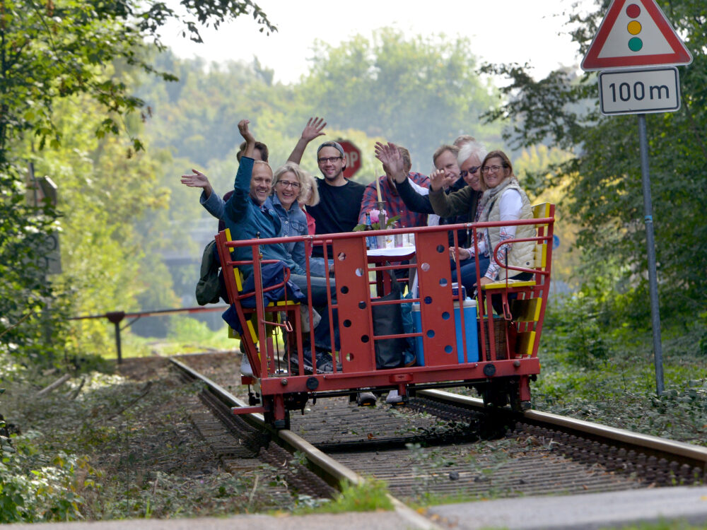 Eine rote Draisine mit mehreren Personen, die auf den Sitzen sitzen und die Hände heben. Im Hintergrund sind Bäume und ein Verkehrsschild mit der Aufschrift '100 m' sichtbar.