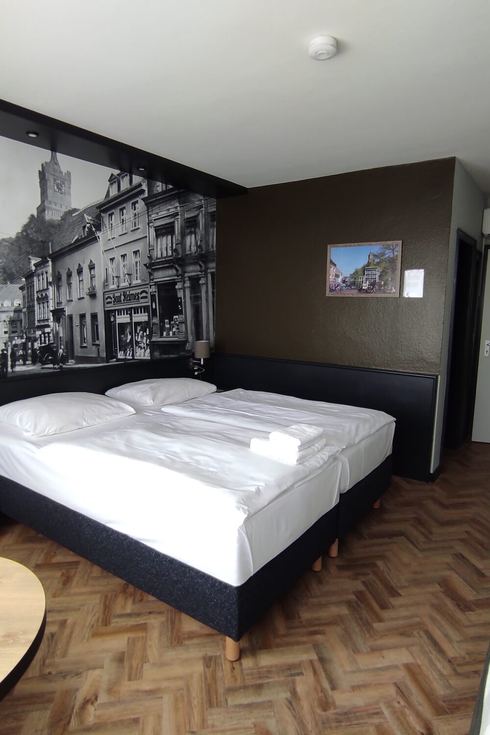 Hotelzimmer im Akzent City Hotel Kleve mit zwei Einzelbetten, Nachttischen mit Lampen, großem schwarz-weiß Foto einer Burg über den Betten, Tisch mit Stuhl und Sessel am Fenster