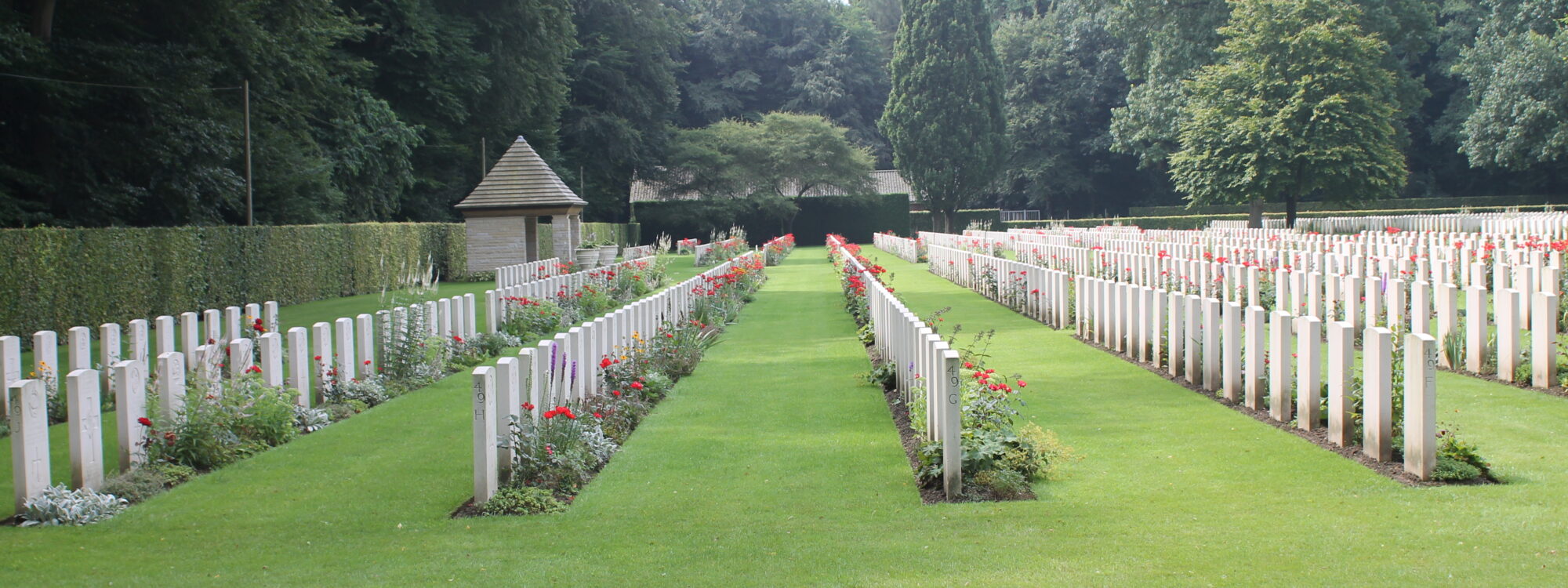 Weiße Grabmale auf dem Britischen Ehrenfriedhof auf einer grünen Wiese, Bäume im Hintergrund