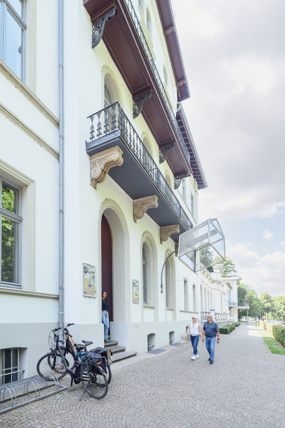 Zwei Personen gehen auf einem gepflasterten Gehweg vor dem Museum Kurhaus Kleve an einem weißen mehrstöckigen Gebäude mit Balkonen und Fahrrädern vorbei, Bäume und Straße rechts sichtbar.