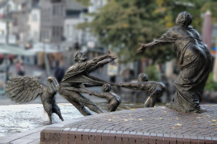 Bronzeskulpturengruppe im Elsabrunnen mit einer Frau, die zwei Kinder an den Händen hält, während ein Engel sie umarmt. Im Hintergrund sind Cafés und Bäume sichtbar.