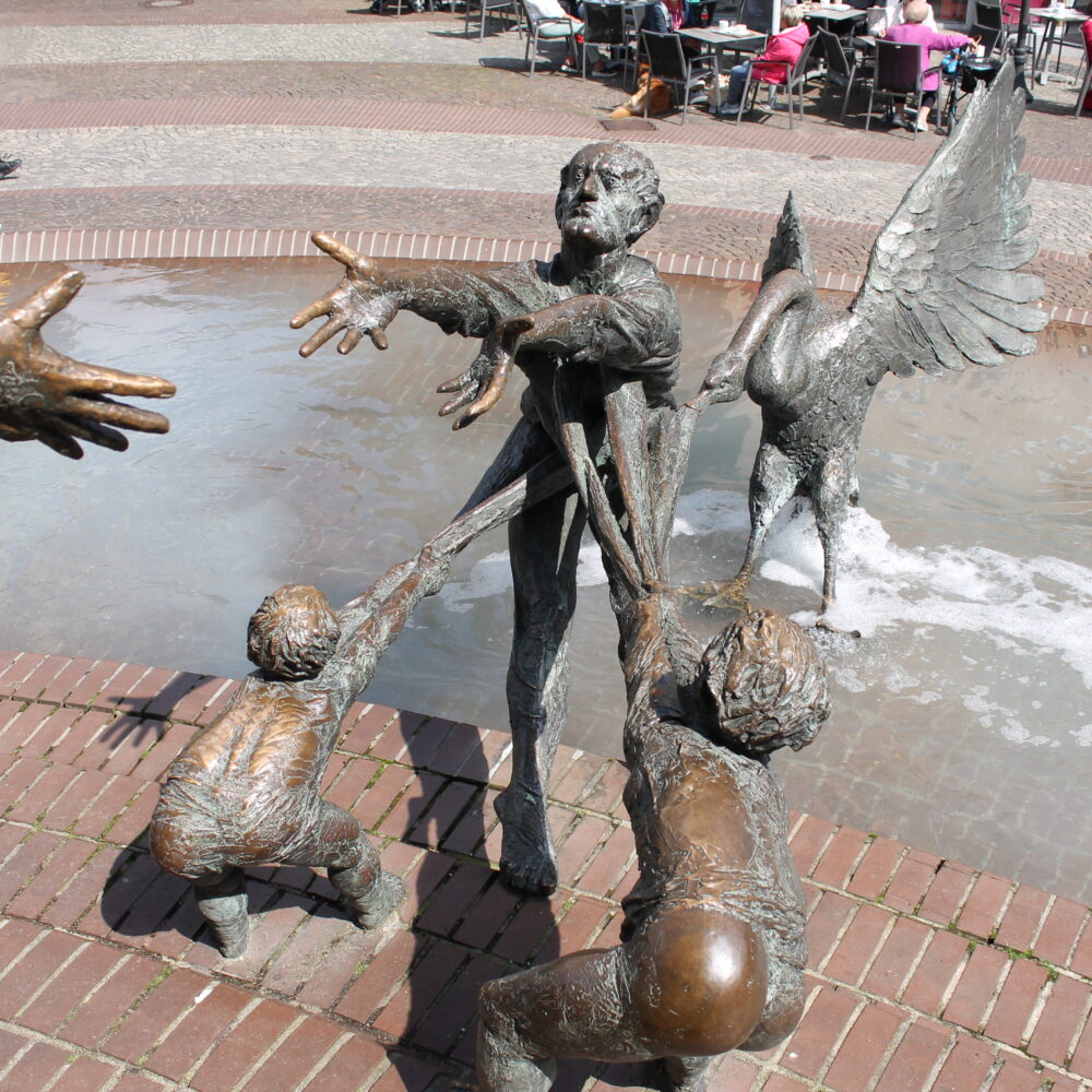 Bronzeskulpturengruppe im Elsabrunnen mit einer Frau, die zwei Kinder an den Händen hält, während ein Engel sie umarmt. Im Hintergrund sind Cafés und Bäume sichtbar.