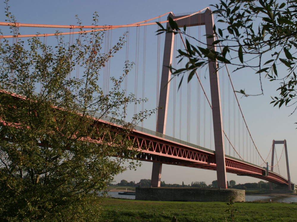Rheinbrücke in Emmerich mit zwei hohen Pylonen und hängenden Stahlseilen, teilweise von Bäumen im Vordergrund verdeckt.