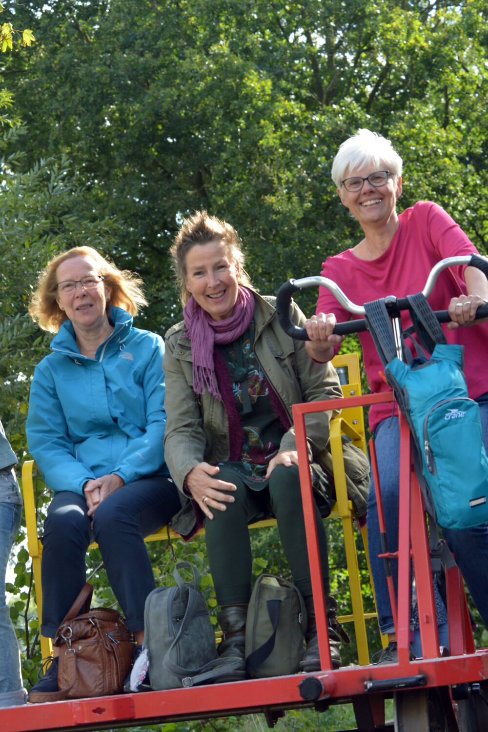 Vier Personen auf Fahrraddraisinen, die auf Schienen fahren. Zwei Frauen sitzen vorne, drei Frauen sitzen hinten. Bäume im Hintergrund.