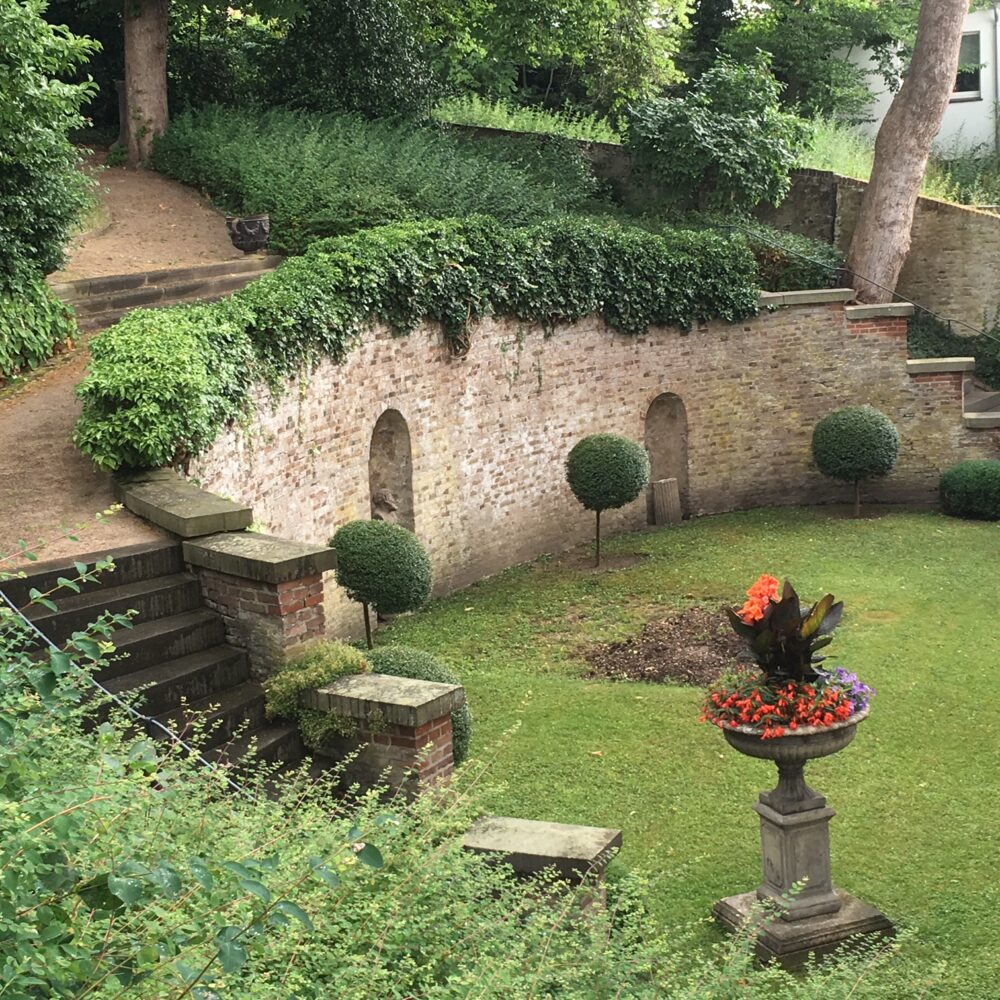 Garten des Museum B.C. Koekkoek-Haus mit gepflegtem Rasen, drei runden Buchsbaumsträuchern, einer hohen Steinmauer mit drei Nischen und zwei großen Pflanzschalen mit roten Blumen.