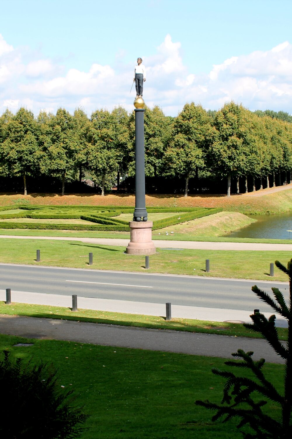 Eiserner Mann auf einem hohen Sockel in den Gartenanlagen, umgeben von einer symmetrischen Anordnung von Bäumen und einem Wasserlauf.