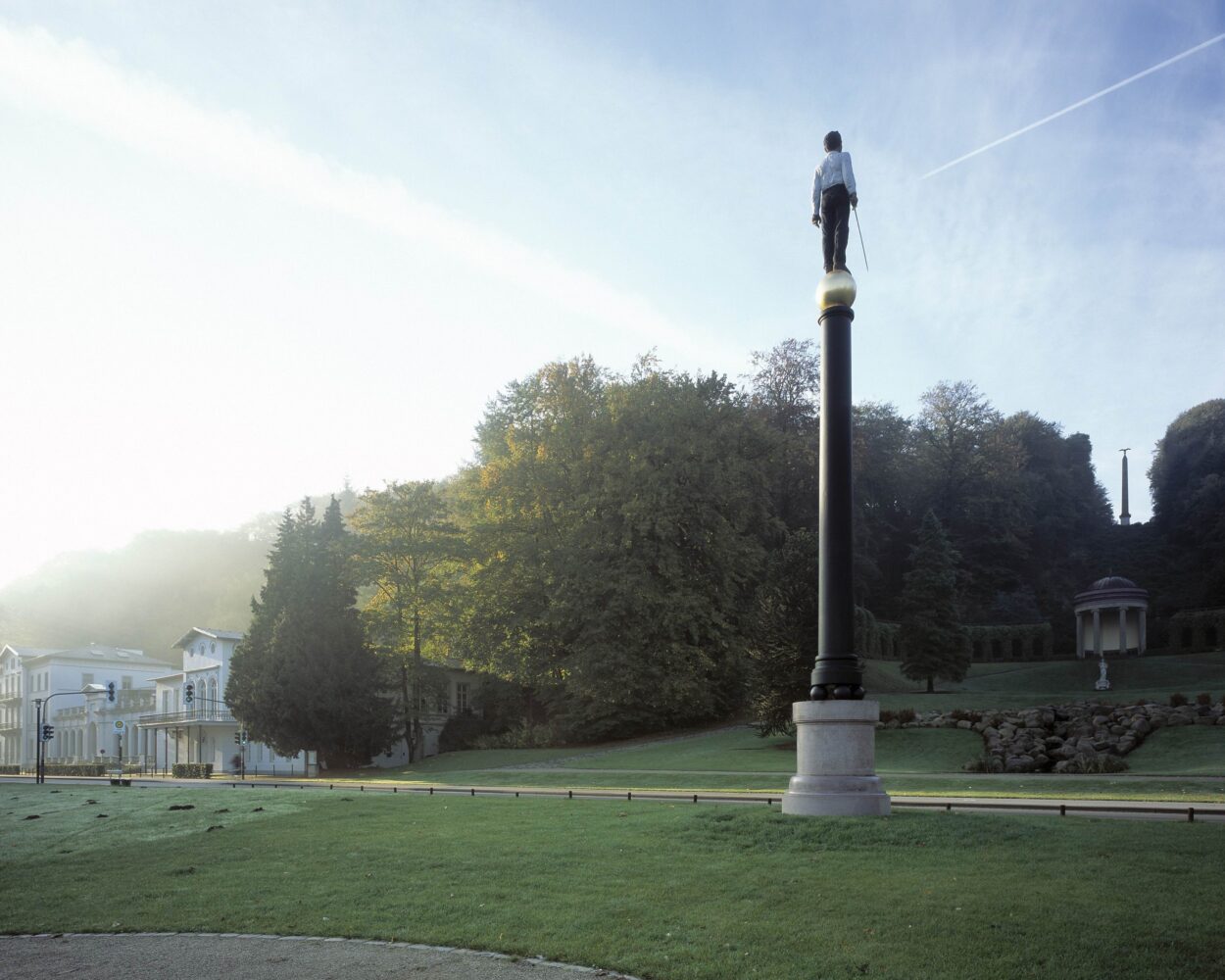 Neuer Eiserner Mann auf einer Säule im Vordergrund, Gartenanlagen mit Museum Kurhaus Kleve und Amphitheater im Hintergrund, Bäume und Rasenflächen.