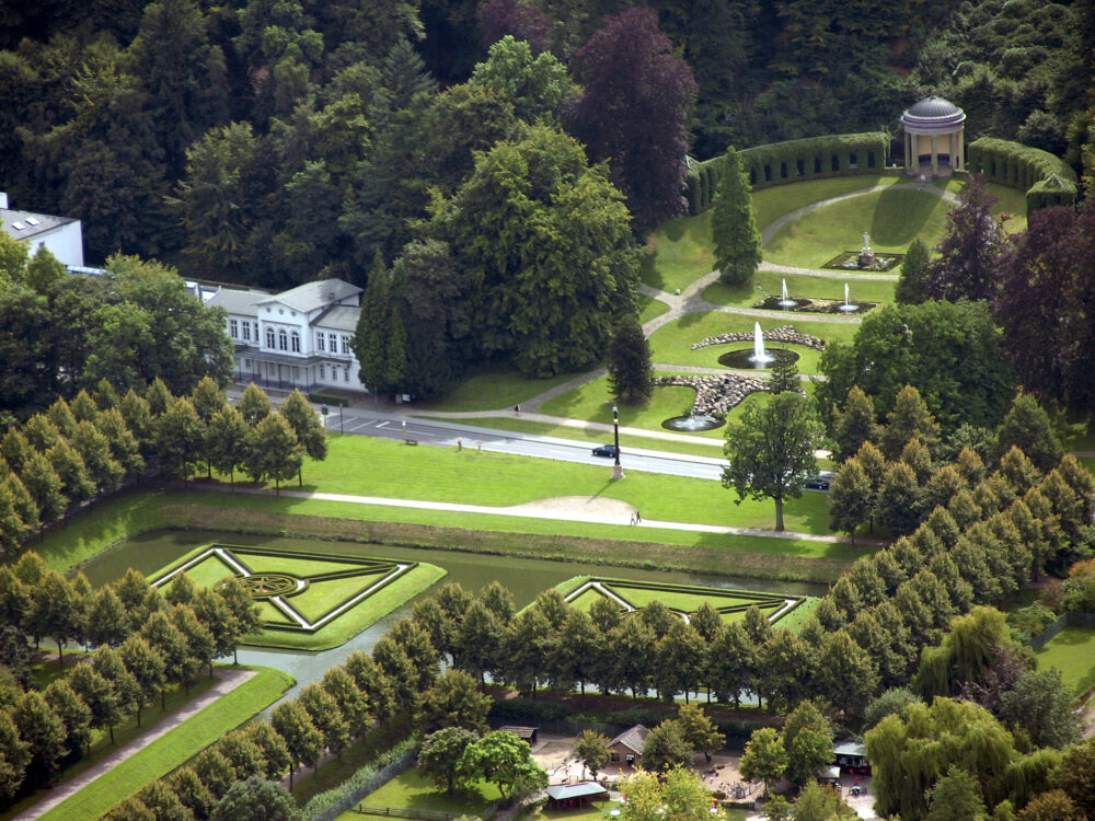 Luftaufnahme der symmetrisch gestalteten Klever Gartenanlage mit Hecken, einem Springbrunnen, einem kleinen Pavillon und von Bäumen umgebenen Wegen.