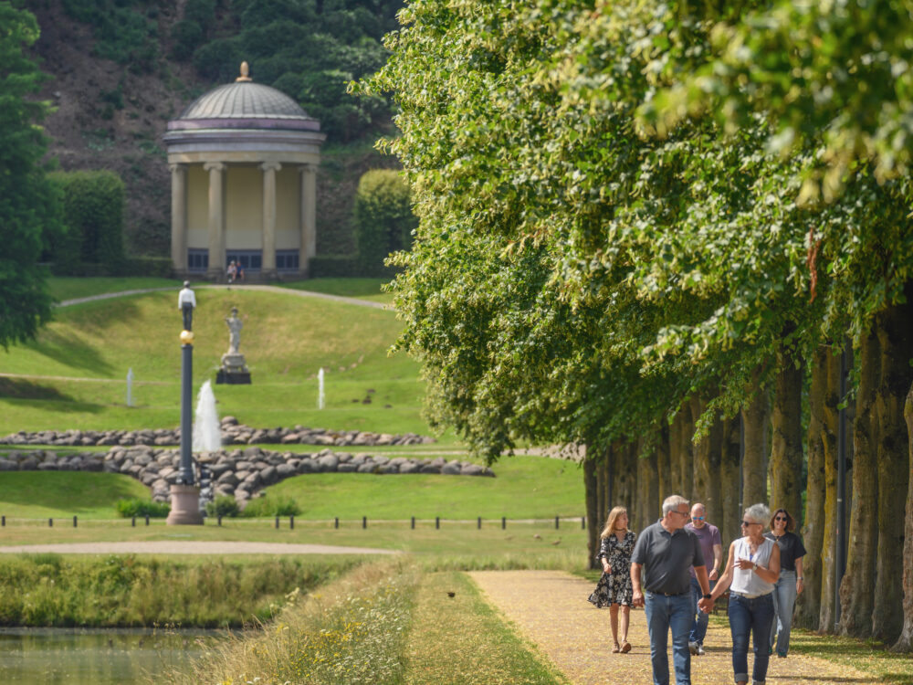 Zwei Spaziergänger gehen auf einem Weg zwischen Bäumen durch die Gartenanlagen. Im Hintergrund steht ein Pavillon und eine Statue, daneben ein Springbrunnen.