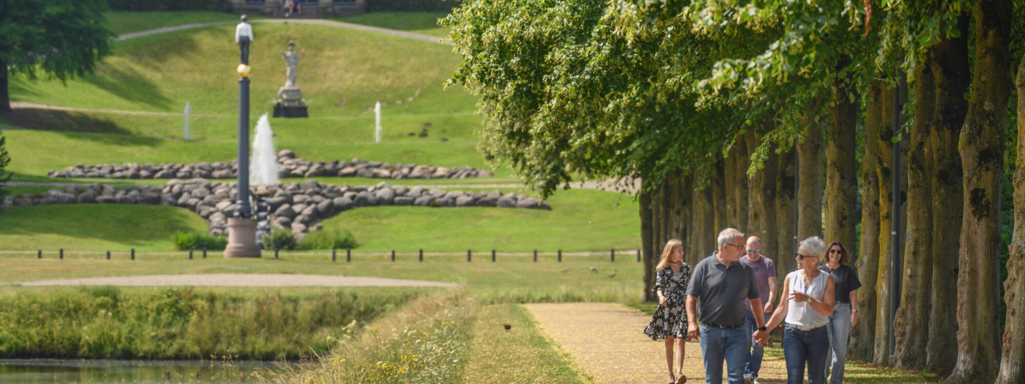Gartenanlagen mit einem Amphitheater im Hintergrund, Spaziergänger auf einem Weg zwischen Bäumen, eine Statue und ein Springbrunnen sichtbar.