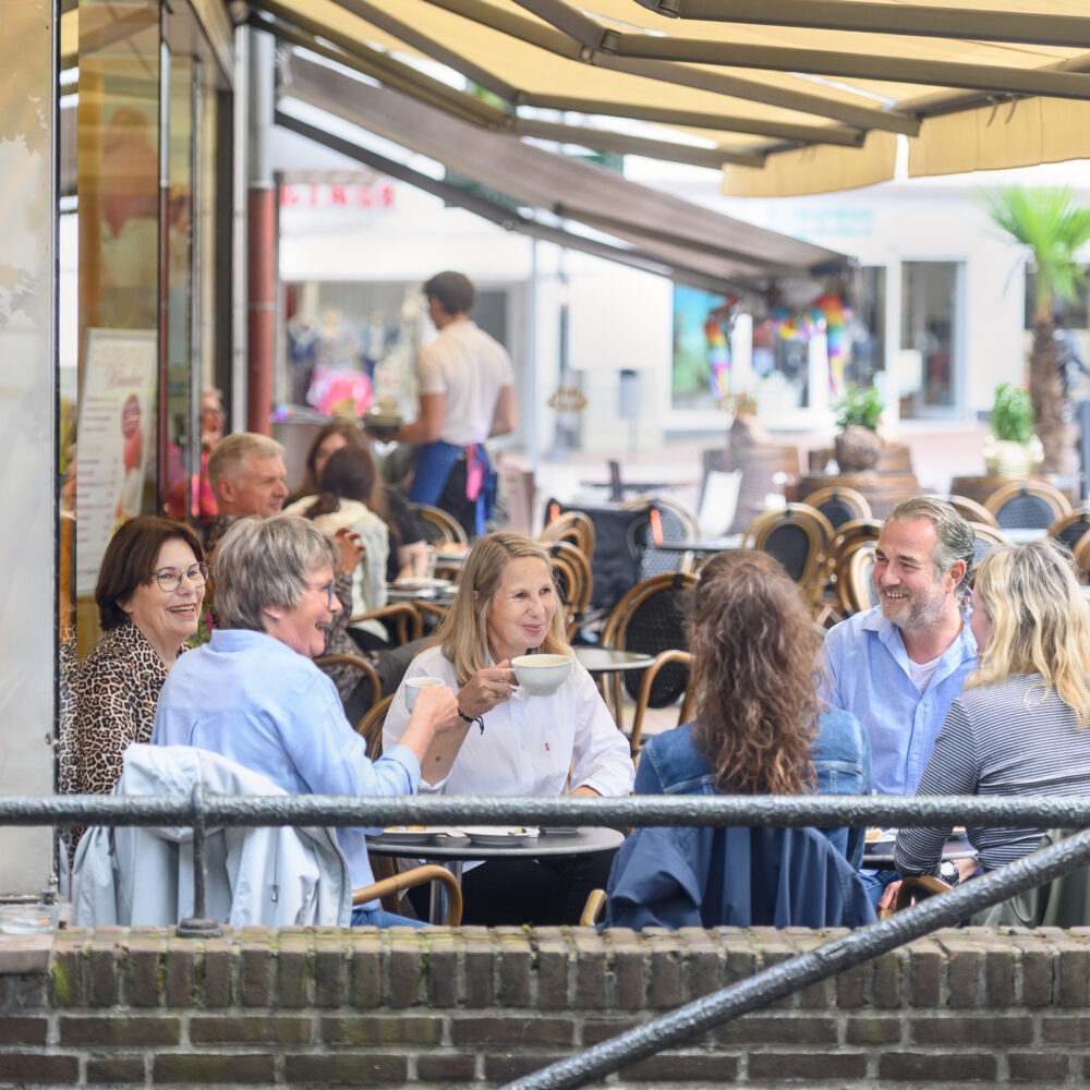 Sechs Personen sitzen in Kleve an zwei Tischen im Außenbereich eines Cafés mit braunen Stühlen und Tischen, umgeben von Bäumen und Gebäuden.