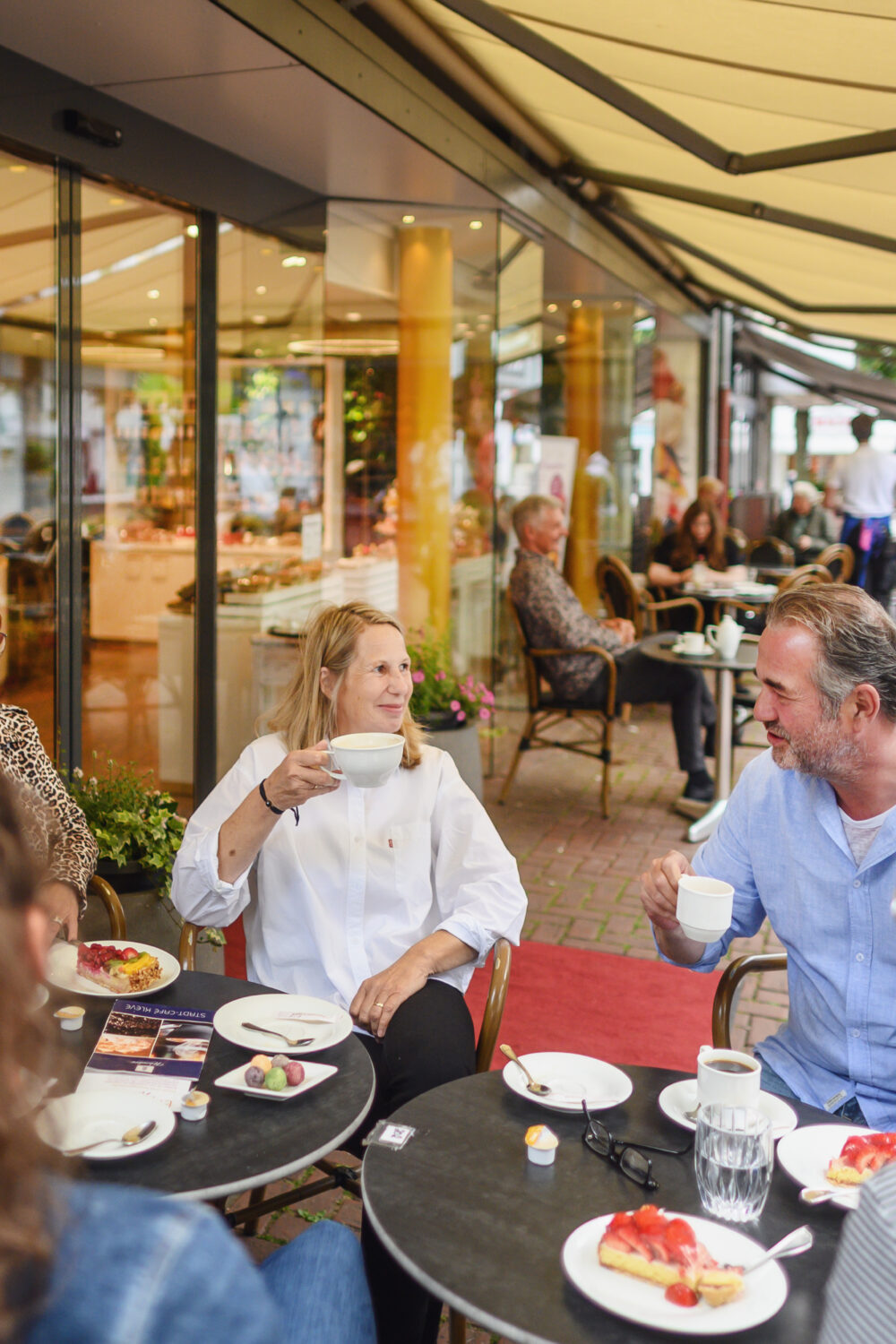 Mehrere Personen sitzen an zwei runden Tischen im Außenbereich eines Cafés, auf den Tischen Teller mit Kuchen und Gläser mit Getränken in Kleve.
