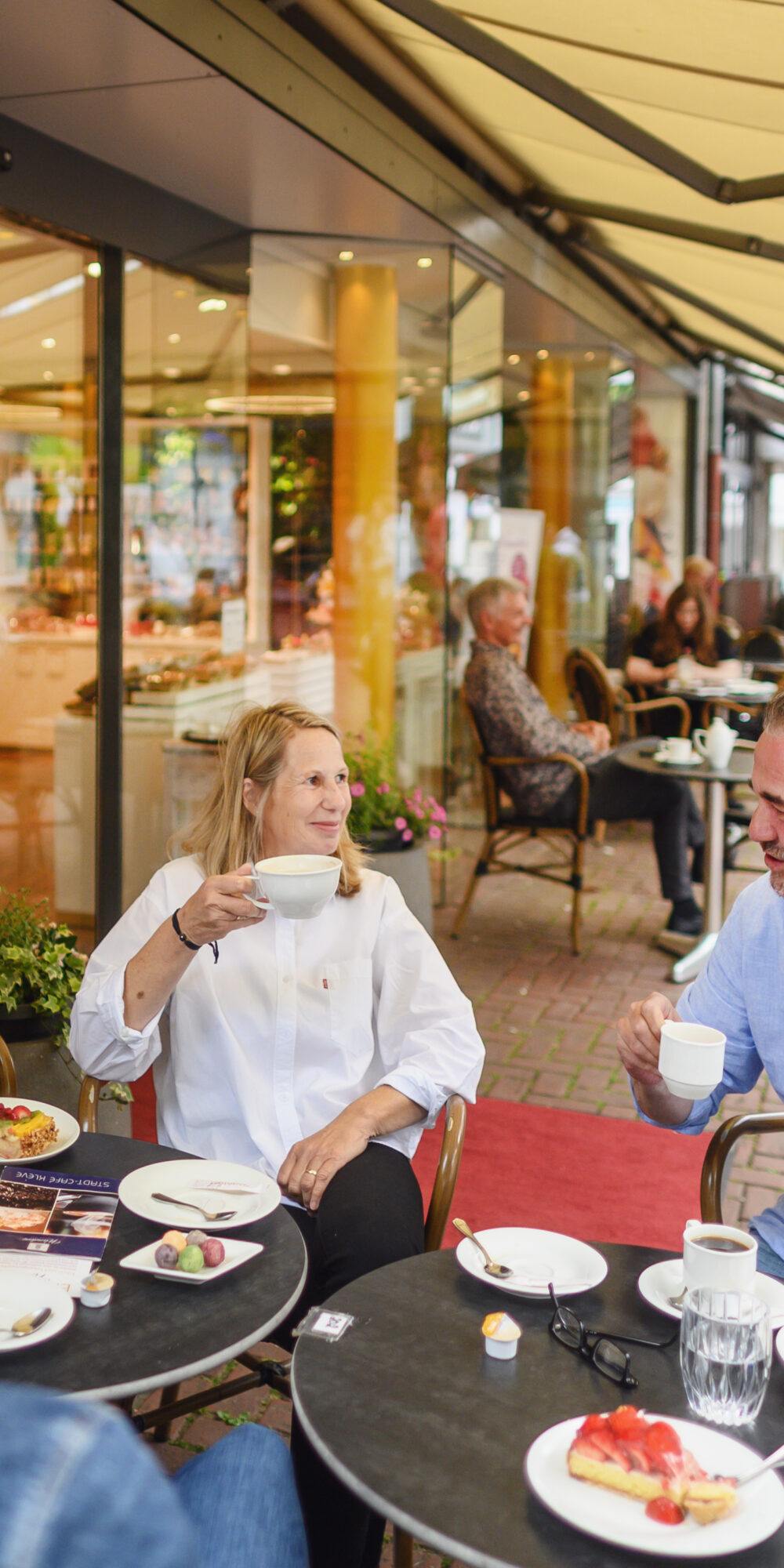Mehrere Personen sitzen an zwei runden Tischen im Außenbereich eines Cafés, auf den Tischen Teller mit Kuchen und Gläser mit Getränken in Kleve.