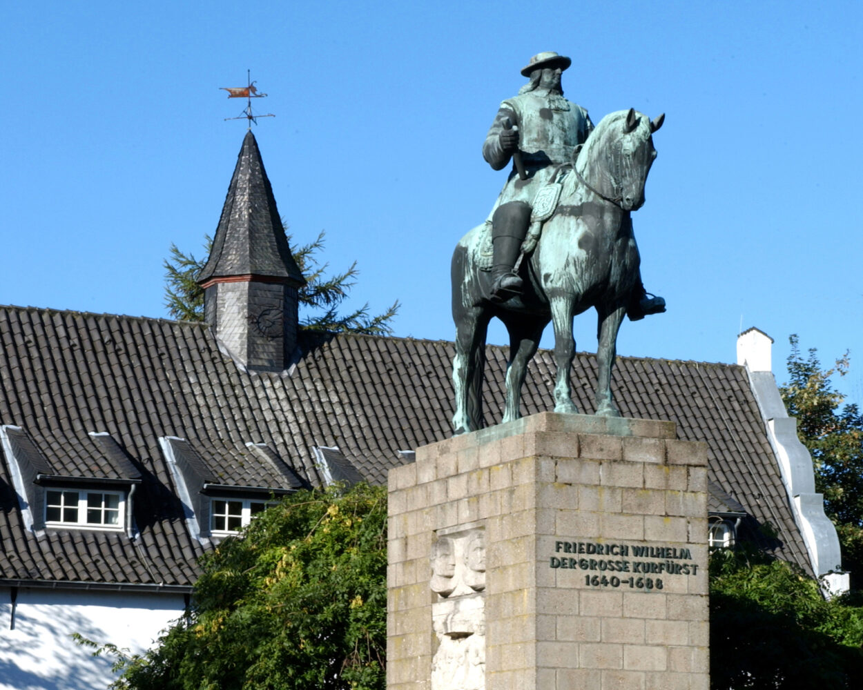 Der Große Kurfürst Friedrich Wilhelm als Bronzestatue auf einem Steinsockel. Er sitzt auf einem Pferd, trägt einen Hut und eine Uniform. Im Hintergrund ein historisches Gebäude mit einem spitzen Dach.