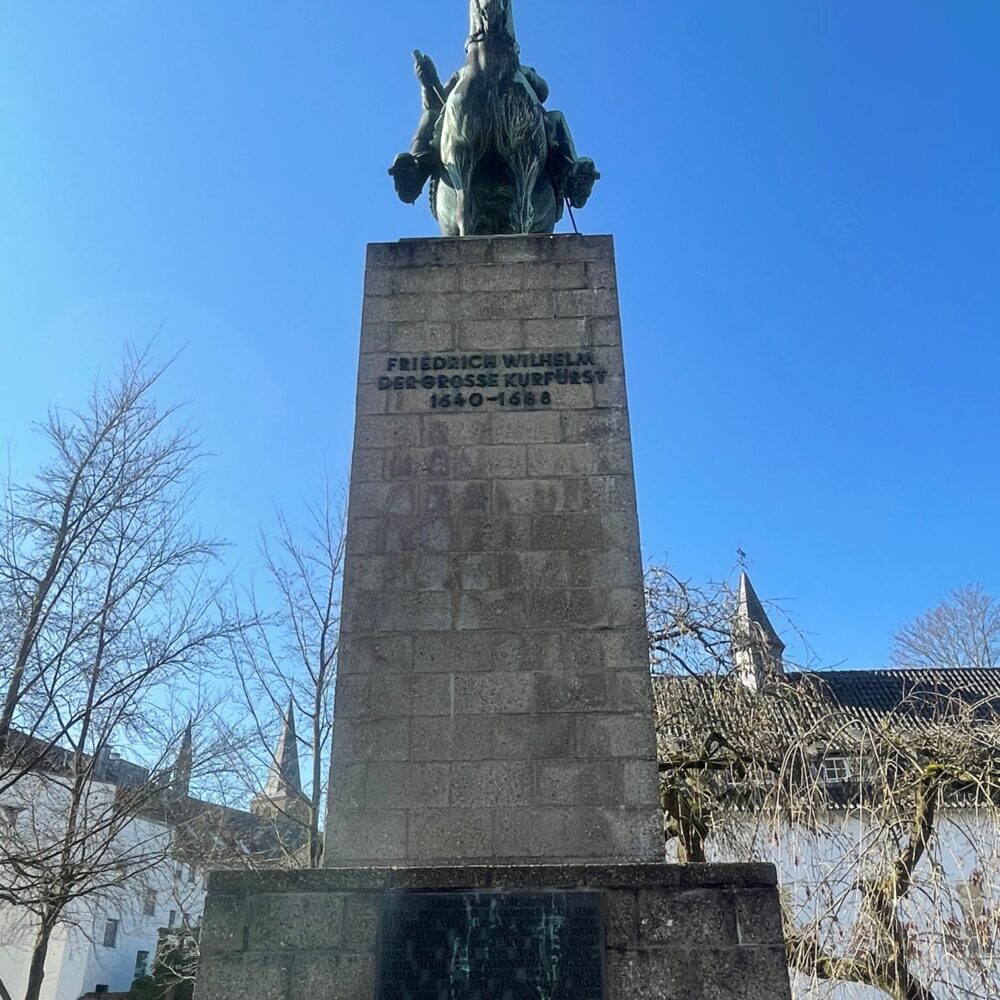 Der Große Kurfürst Friedrich Wilhelm als Bronzestatue auf einem Steinsockel. Er sitzt auf einem Pferd, trägt einen Hut und eine Uniform. Im Hintergrund Bäume. Er sitzt auf einem Pferd, trägt einen Hut und eine Uniform. Im Hintergrund ein historisches Gebäude mit einem spitzen Dach.