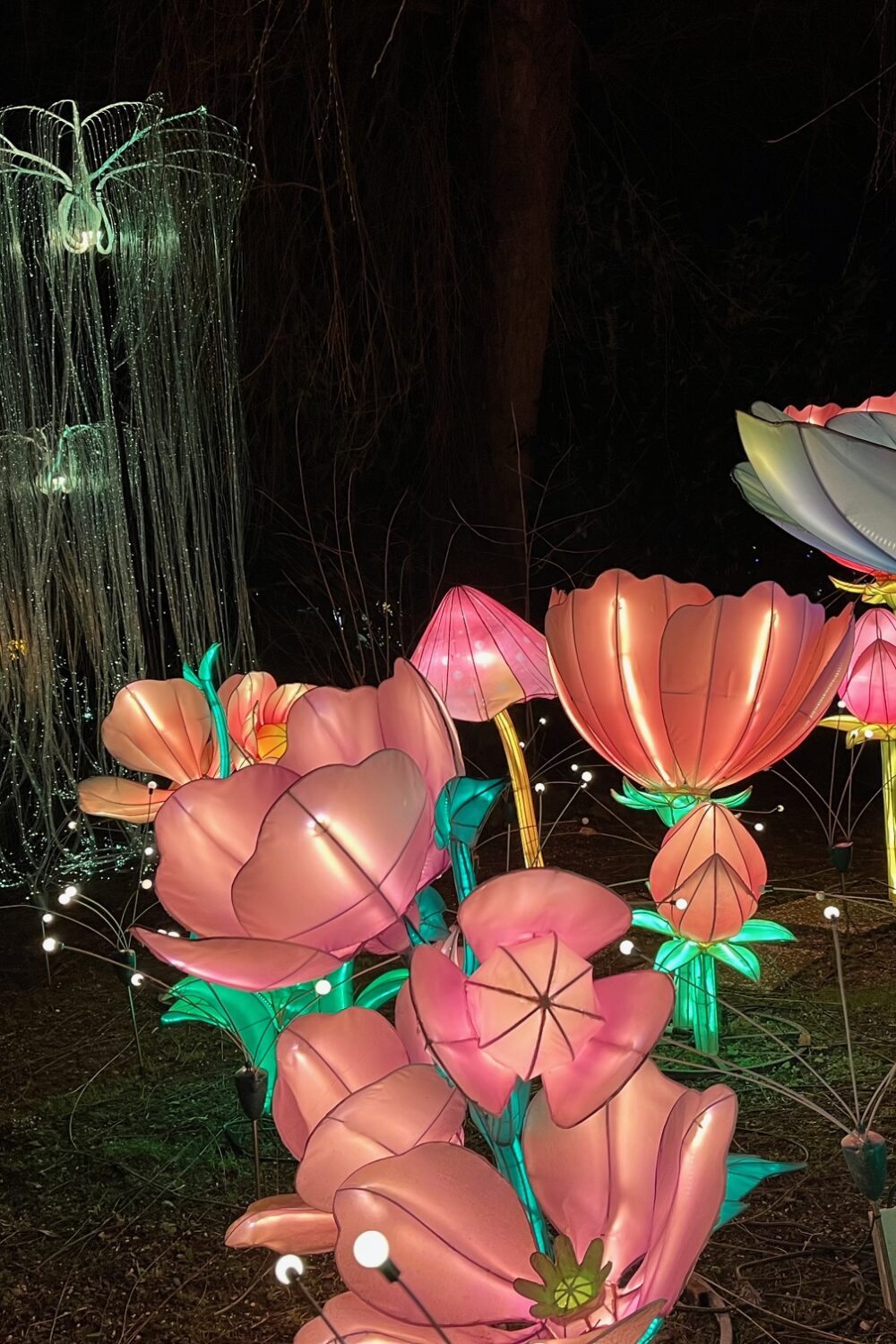 Bunte, leuchtende Blumen aus Kunststoff in verschiedenen Formen und Größen, darunter große rosa und weiße Blüten, vor einem dunklen Hintergrund. Im Hintergrund sind leuchtende Quallen und weitere Blumen sichtbar.