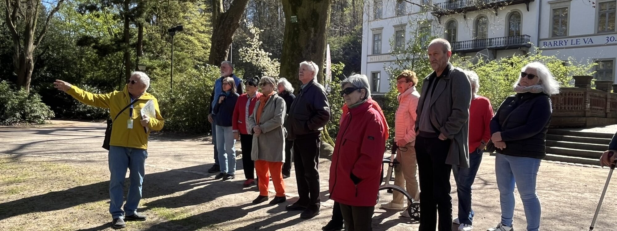 Eine Gruppe von Personen steht auf einem Weg in einem Park. Eine Person zeigt mit dem Finger in eine Richtung. Im Hintergrund ist ein Gebäude mit mehreren Fenstern und einem Balkon sichtbar.