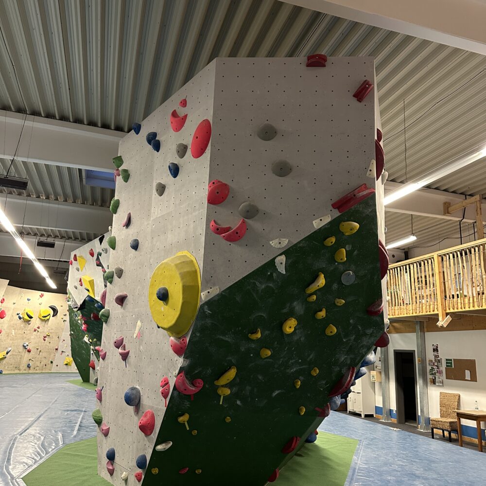 Boulderwand in einer Kletterhalle, dreieckige Form mit bunten Griffen in Rot, Blau, Gelb und Grün, grüne Unterlage, Holzgeländer im Hintergrund.