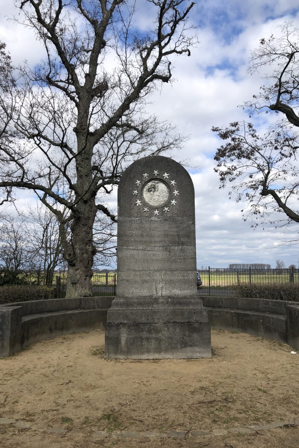 Johanna-Sebus-Denkmal, graue Stele mit einer rechteckigen Tafel in der Mitte, umgeben von einer runden, niedrigen Mauer, umgeben von Bäumen.