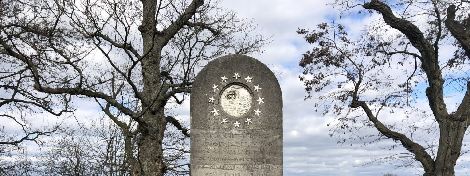 Denkmal für Johanna Sebus, grauer Stein mit einer runden Medaille und Sternen, umgeben von einer halbkreisförmigen Bank aus Beton, Bäume im Hintergrund.
