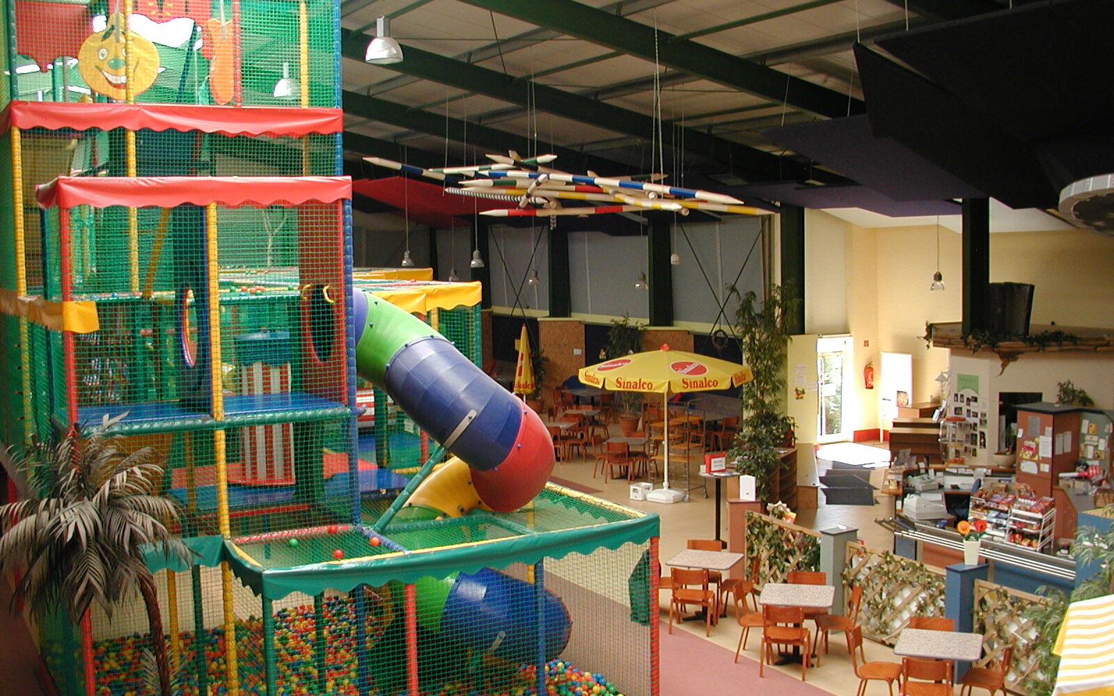 Blick auf ein Klettergerüst in einem Spielplatz mit bunten Bällen, Rutsche und Netz. Im Hintergrund sind Tische und Stühle sowie ein Essensbereich sichtbar.