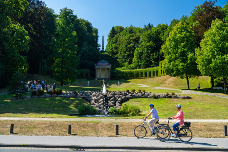 Zwei Radfahrer auf einem Weg im Kleve, im Hintergrund eine Gruppe von Menschen vor einem Amphitheater und einer Wasserfontäne, umgeben von Bäumen.
