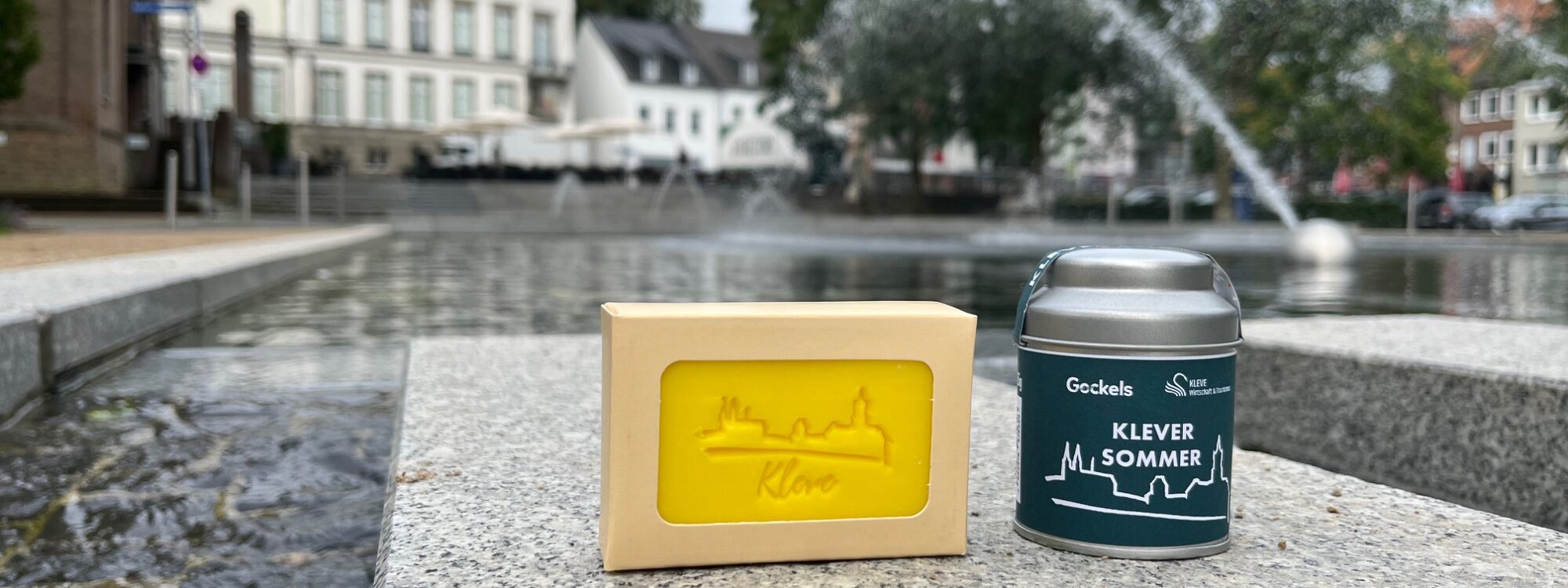 Klever Souvenirs: Gelbe Seife mit Stadt-Silhouette und Dose mit Aufschrift 'Klever Sommer' auf grauem Stein am Wasserbecken vor Bäumen und Gebäuden