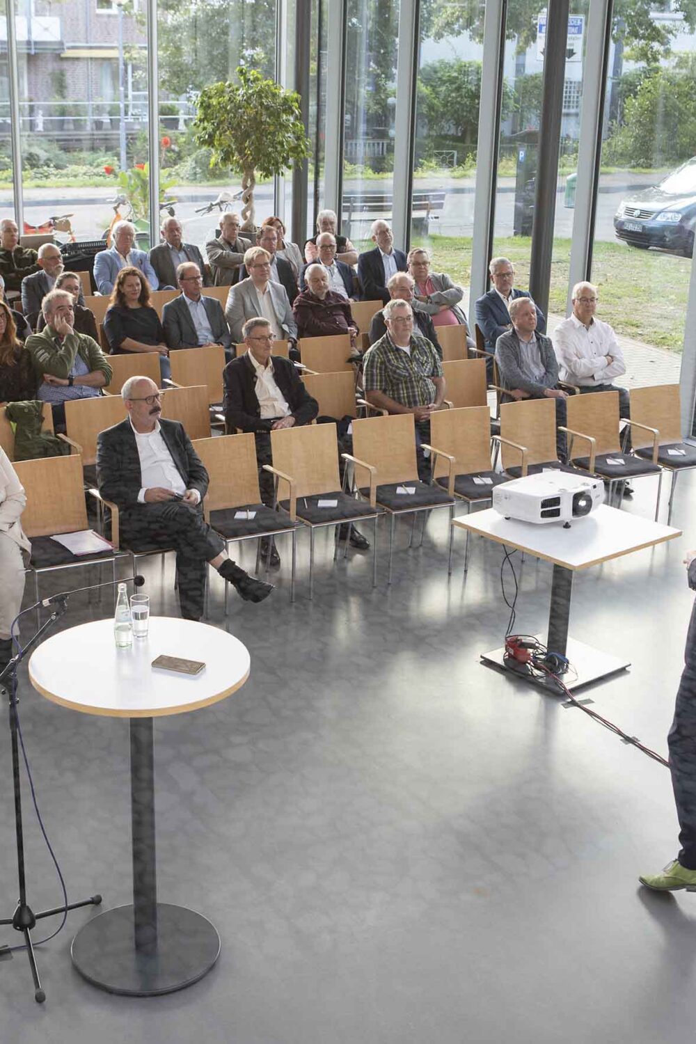 Ein Mann hält einen Vortrag vor einem Publikum in einem hellen Raum mit großen Fenstern in der Stadthalle Kleve und Blick auf eine Straße mit parkenden Autos.