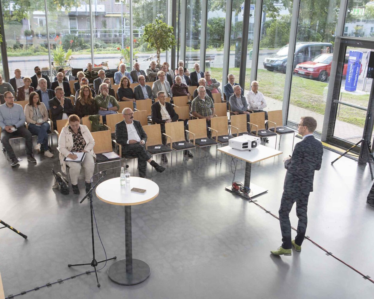 Ein Mann hält einen Vortrag vor einem Publikum in einem hellen Raum mit großen Fenstern in der Stadthalle Kleve und Blick auf eine Straße mit parkenden Autos.