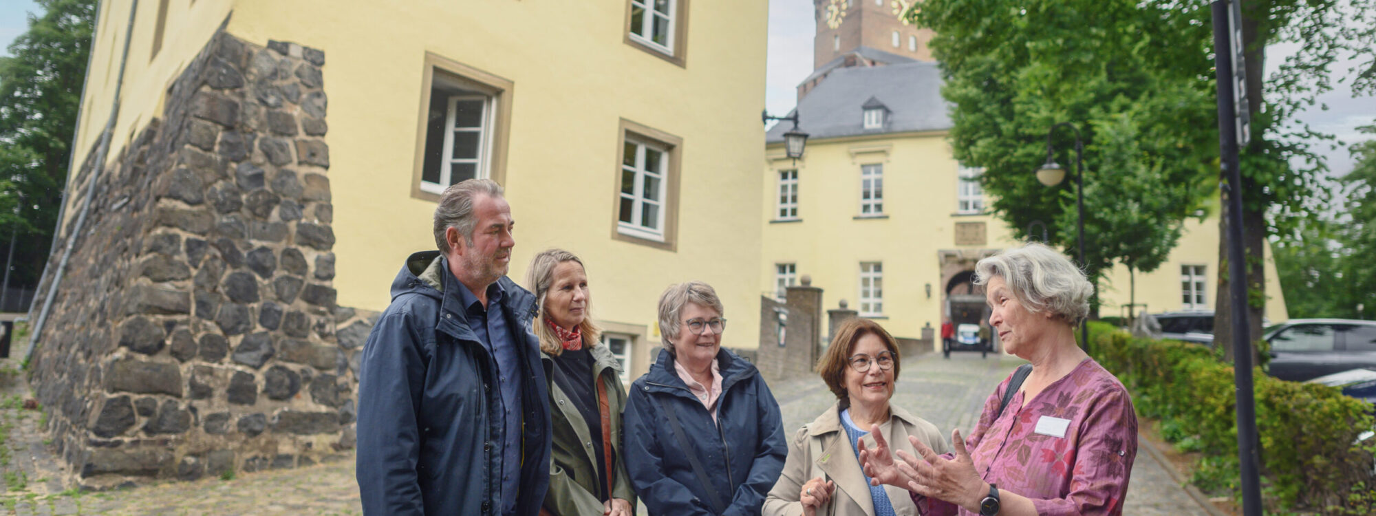 Fünf Personen stehen auf einem gepflasterten Weg vor einem gelben Gebäude mit einer Steinmauer. Im Hintergrund ist die Schwanenburg sichtbar. Zur erkennen ist eine Gästeführerin die Stadtführungen in Kleve anbietet.