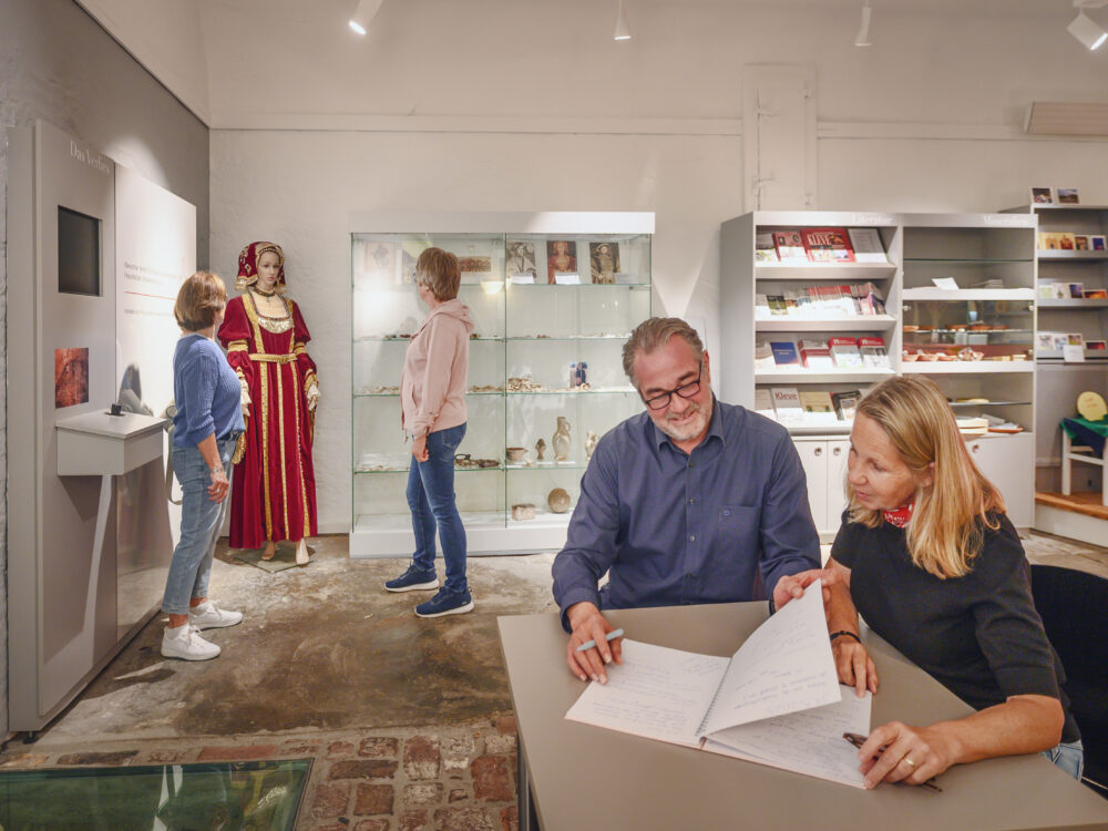Drei Personen stehen in einem Ausstellungsraum, eine in einem historischen Kostüm. Ein Tisch mit Notizen und einem Laptop ist im Vordergrund.