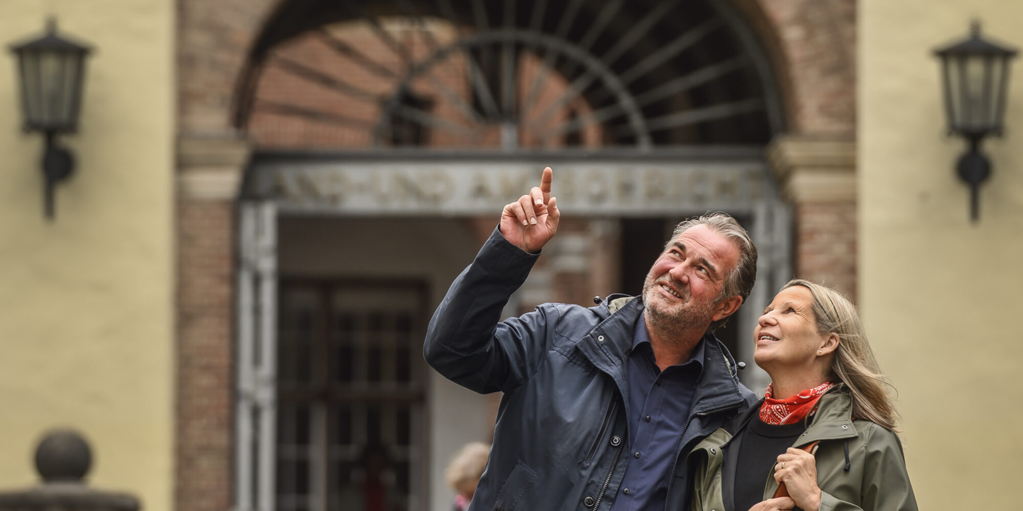 Zwei Personen stehen vor einem historischen Torbogen mit Laternen an der Schwanenburg an den Seiten, eine Person zeigt nach oben.