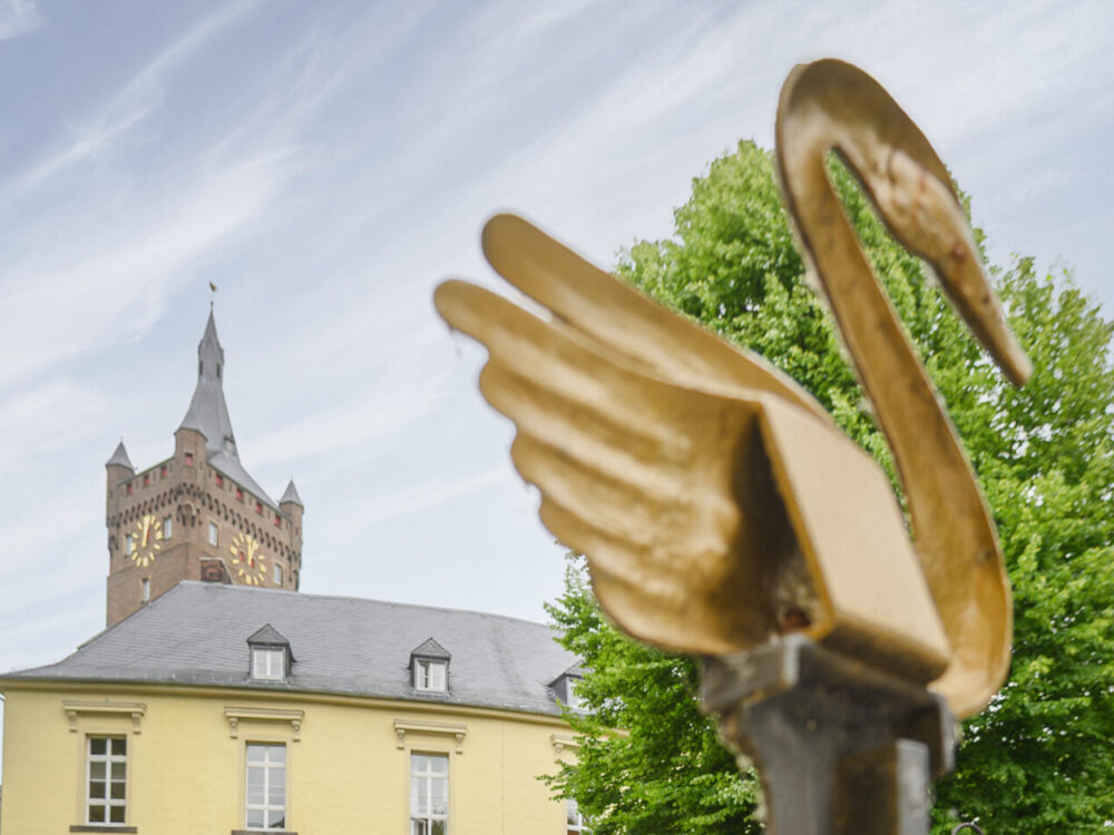 Zwei Personen stehen vor einem gelben Gebäude mit einem Turm im Hintergrund, der Schwanenburg. Im Vordergrund ist ein metallischer Schwan mit ausgebreiteten Flügeln zu sehen.