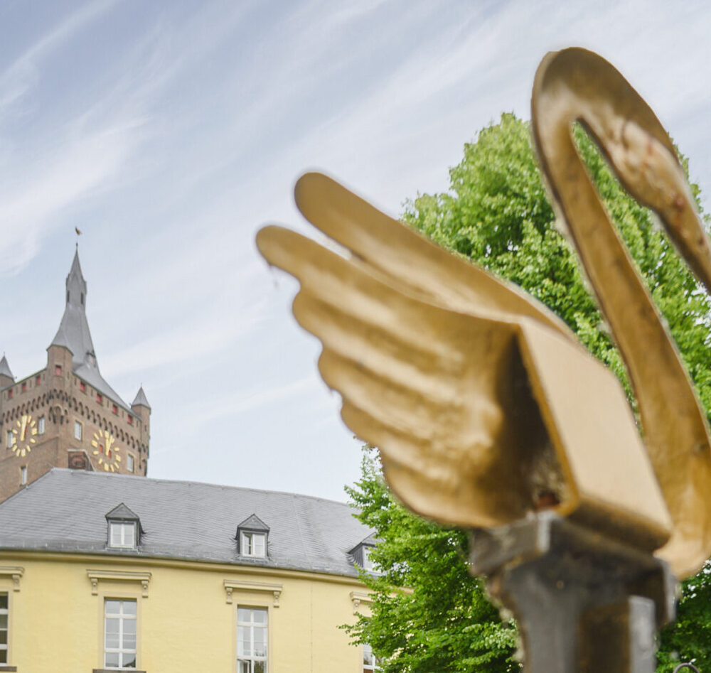 Zwei Personen stehen vor einem gelben Gebäude mit einem Turm im Hintergrund, der Schwanenburg. Im Vordergrund ist ein metallischer Schwan mit ausgebreiteten Flügeln zu sehen.