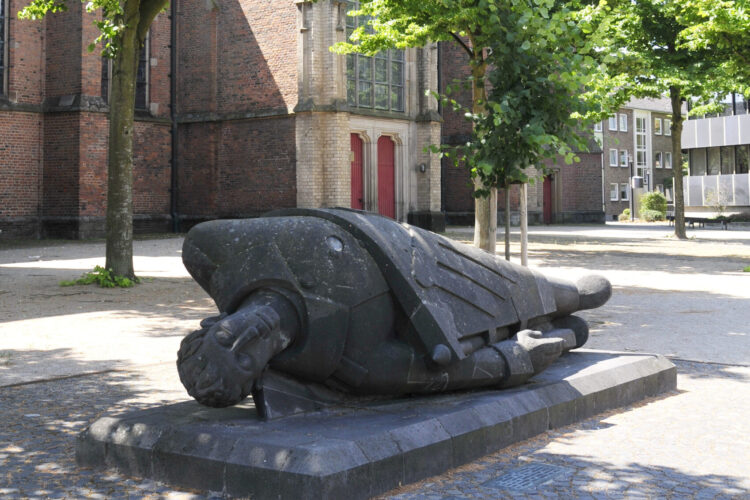 Skulptur eines toten Kriegers, liegend auf einem Sockel, umgeben von Bäumen und einer Kirche im Hintergrund. Der Krieger trägt eine Rüstung und hat einen ernsten Gesichtsausdruck.