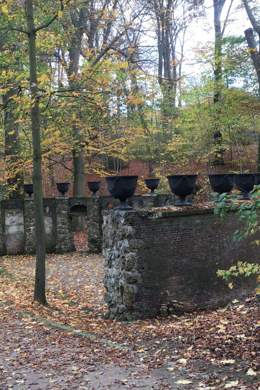 Zwei alte, teilweise verfallene Mauern mit mehreren schwarzen Behältern auf der Oberseite, umgeben von herbstlichen Bäumen mit buntem Laub.