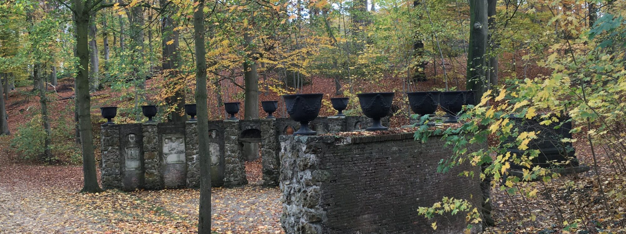 Zwei alte, teilweise verfallene Mauern mit mehreren schwarzen Behältern auf der Oberseite, umgeben von herbstlichen Bäumen mit buntem Laub.
