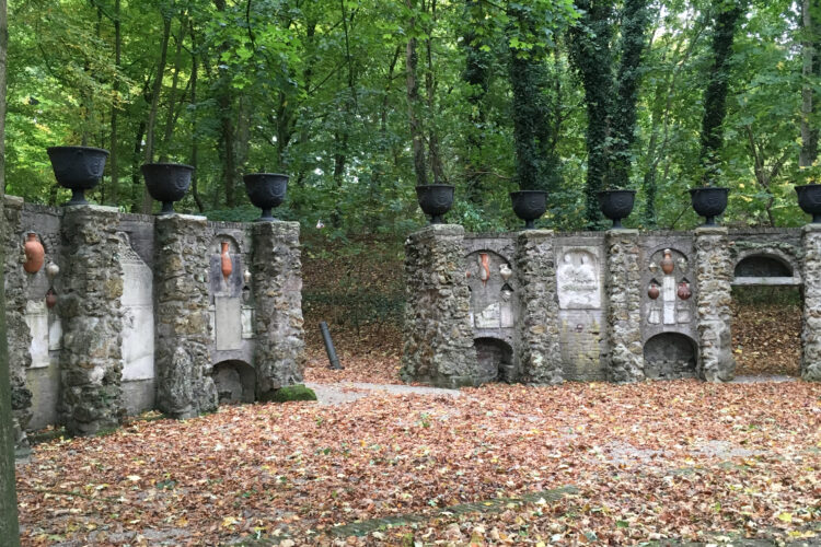 Zwei alte, teilweise verfallene Mauern mit mehreren schwarzen Behältern auf der Oberseite, umgeben von herbstlichen Bäumen mit buntem Laub.