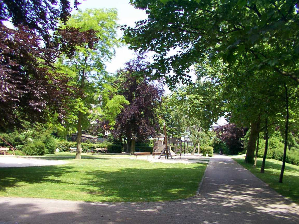 Blick auf einen Park mit Rasenflächen, Bäumen und einem Spielplatz im Hintergrund. Der Weg verläuft rechts im Bild.