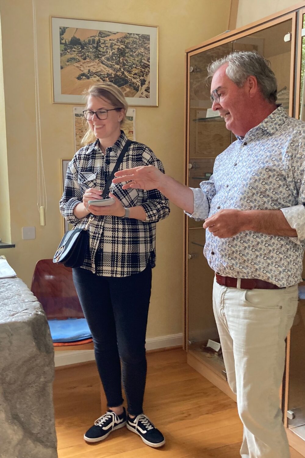 Zwei Personen stehen im Museum Forum Arenacum vor der Nachbildung des Mars Camulus Stein und einer Vitrine mit antiken Artefakten, an der Wand hängt ein gerahmtes Bild.