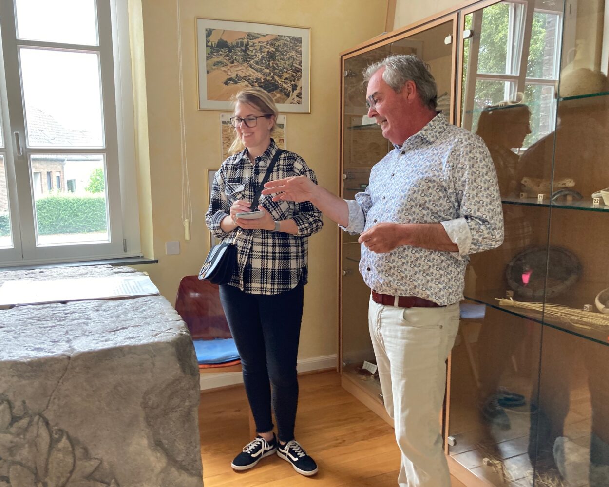 Zwei Personen stehen im Museum Forum Arenacum vor der Nachbildung des Mars Camulus Stein und einer Vitrine mit antiken Artefakten, an der Wand hängt ein gerahmtes Bild.