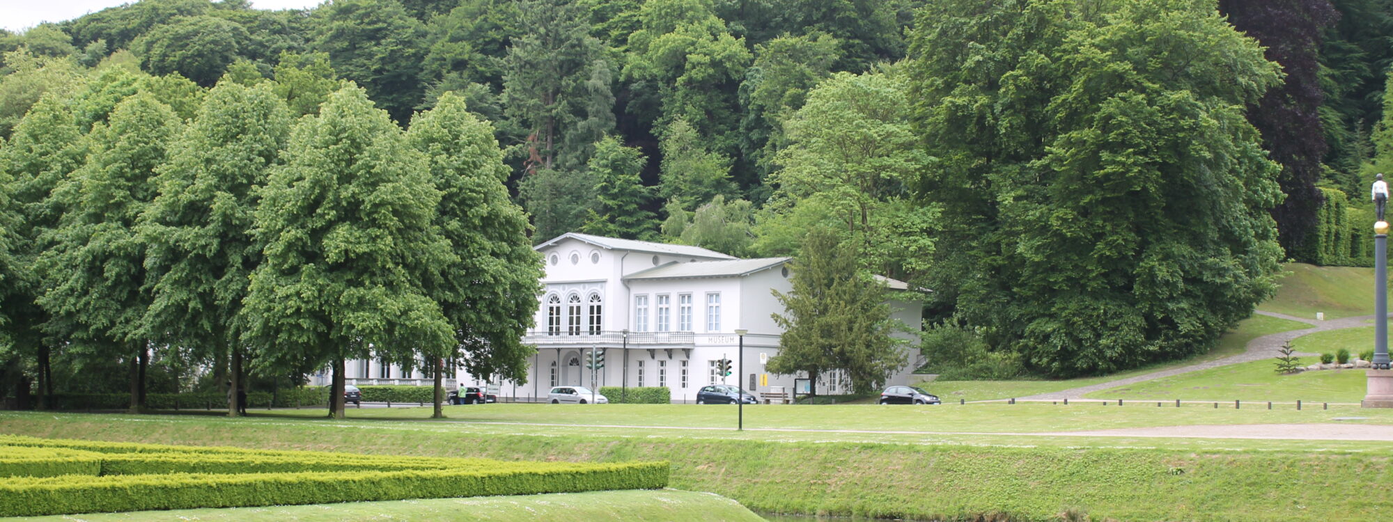 Weißes klassizistisches Gebäude des Museum Kurhaus Kleve vor dichtem Wald, davor ein Teich mit grünen, geometrisch geschnittenen Hecken und Rasenflächen der historischen Gartenanlagen.