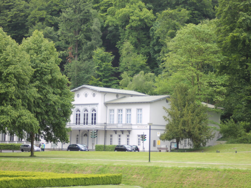 Weißes Gebäude des Museums Kurhaus Kleve mit mehreren Fenstern, umgeben von grünen Bäumen und Rasenflächen der historischen Gartenanlagen.