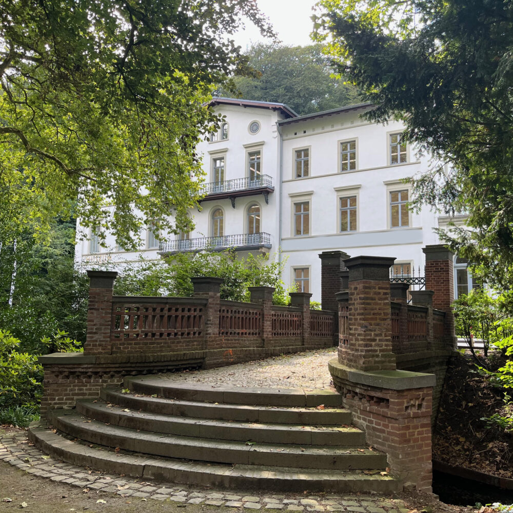 Weißes dreistöckiges Gebäude des Museum Kurhaus Kleve mit Balkonen, umgeben von Bäumen, davor eine breite Treppe mit Brücke und Ziegelgeländer.