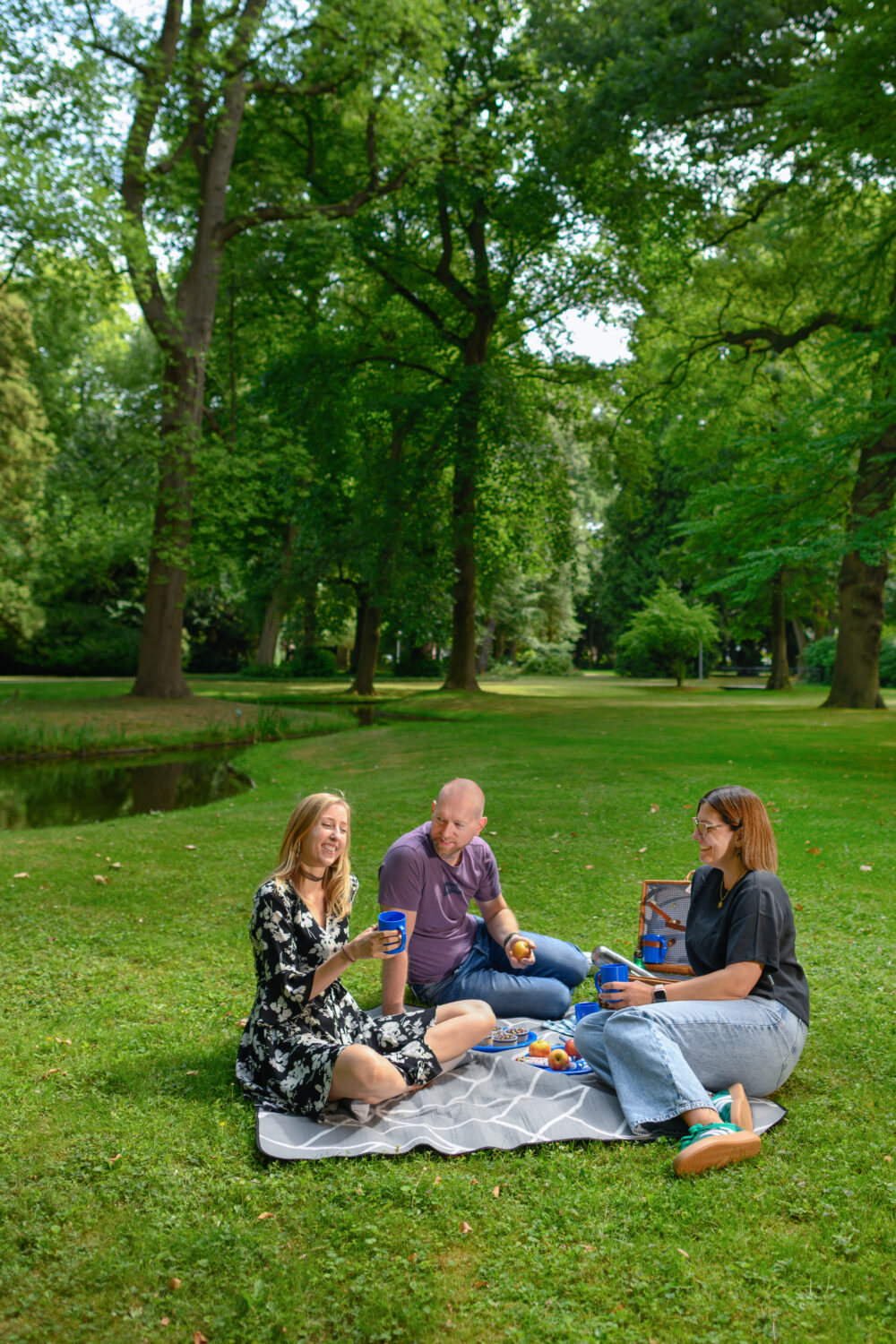 Drei Personen sitzen auf einer Decke im Forstgarten in Kleve auf einer Wiese in einem Park, umgeben von großen Bäumen und einem kleinen Teich.