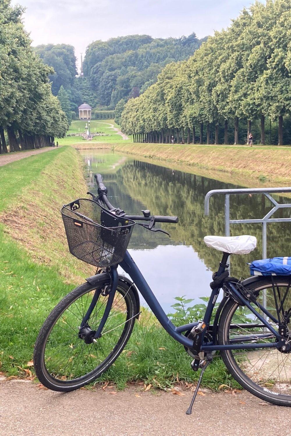 Schwarzes Fahrrad mit Korb steht auf einem Weg neben einem Kanal, umgeben von Bäumen und einer Gartenanlage im Hintergrund.