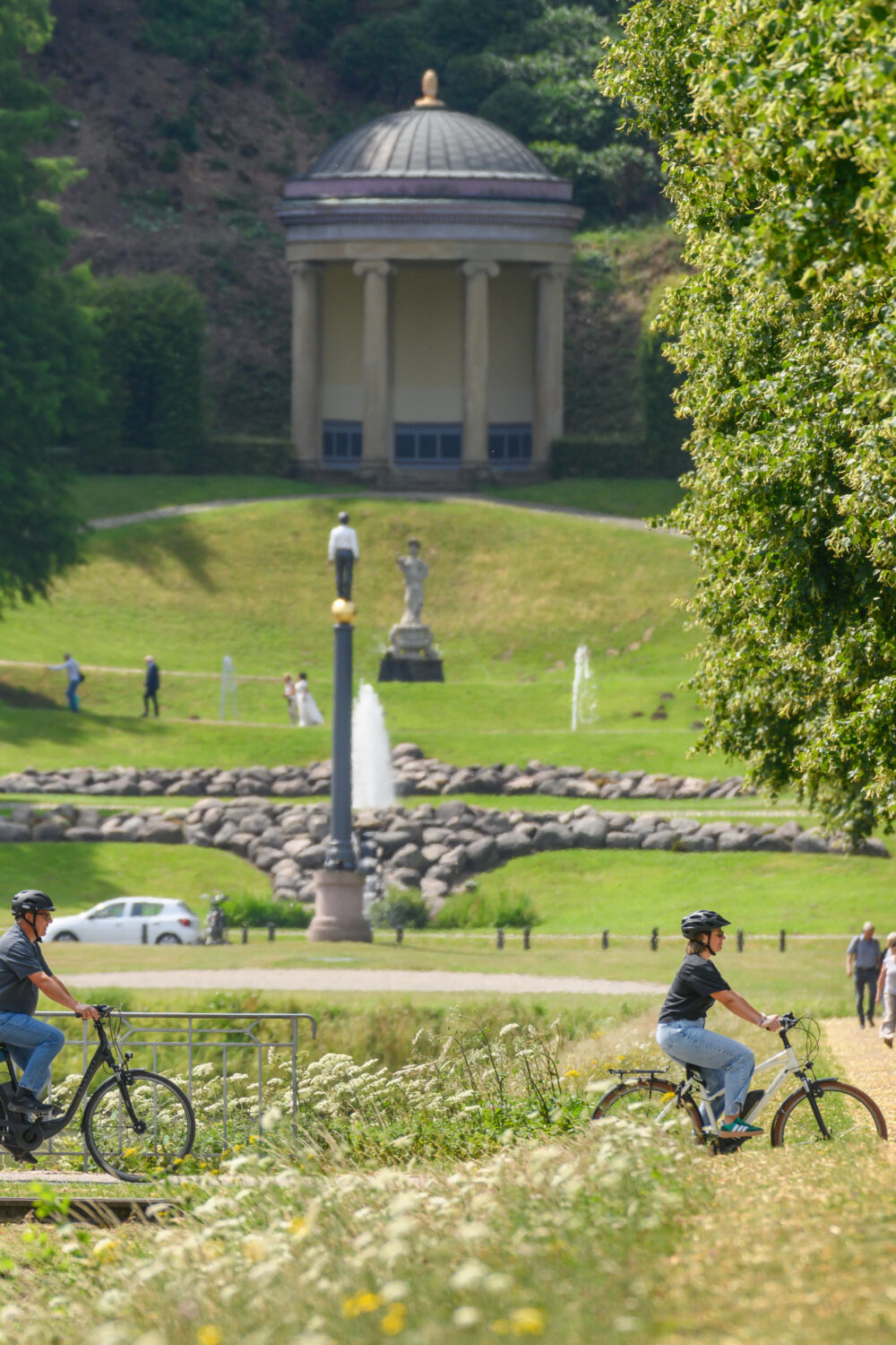 Vier Radfahrer fahren auf einem von Bäumen gesäumten Weg in einem Park, im Hintergrund ein klassizistischer Pavillon mit Säulen und Kuppeldach.