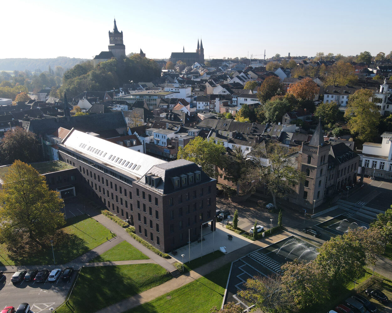 Luftaufnahme von Kleve mit dem modernen Rathausgebäude im Vordergrund inklusive der Tourist Information und der Schwanenburg auf einem Hügel im Hintergrund, umgeben von Wohnhäusern und Bäumen.