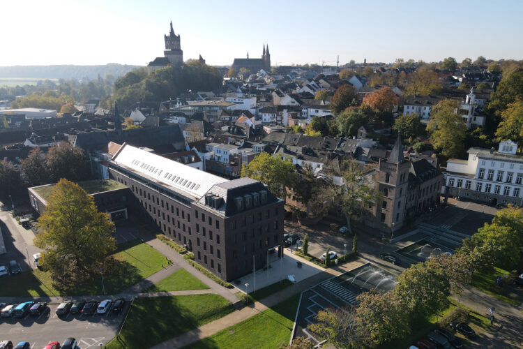 Luftaufnahme von Kleve mit dem modernen Rathausgebäude im Vordergrund inklusive der Tourist Information und der Schwanenburg auf einem Hügel im Hintergrund, umgeben von Wohnhäusern und Bäumen.