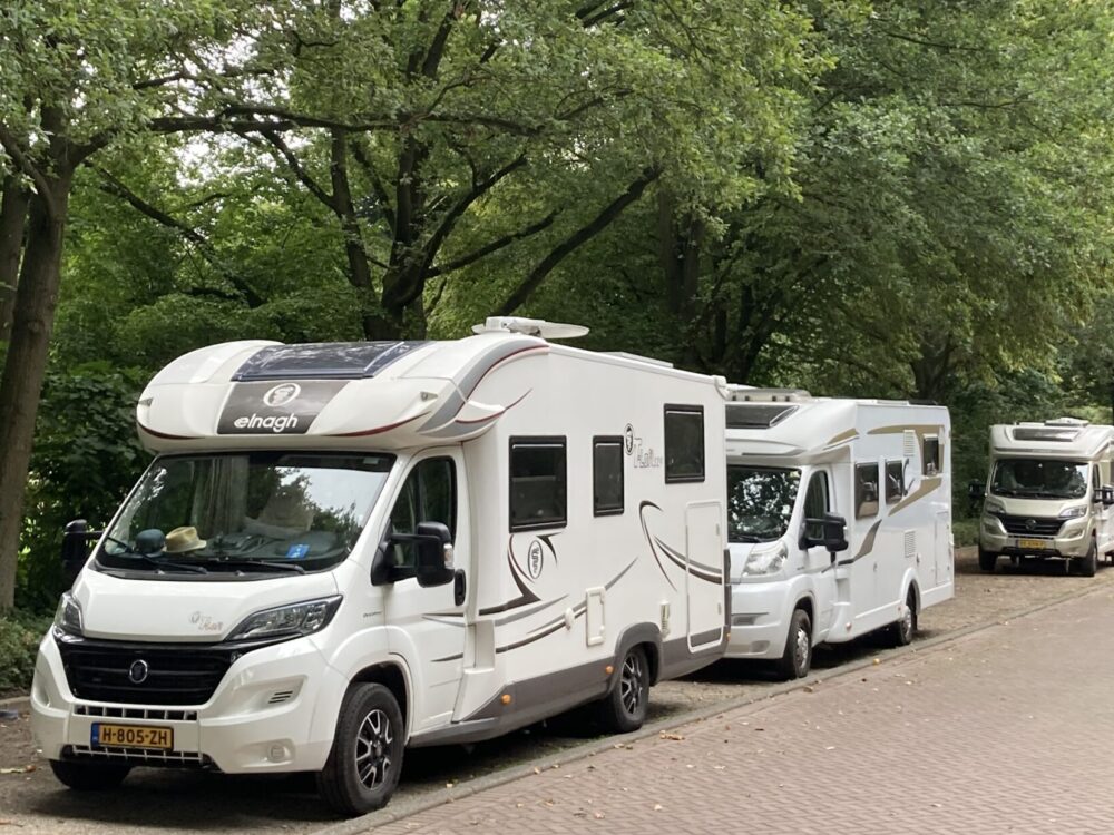 Vier weiße Reisemobile stehen hintereinander auf einem gepflasterten Stellplatz unter grünen Bäumen.